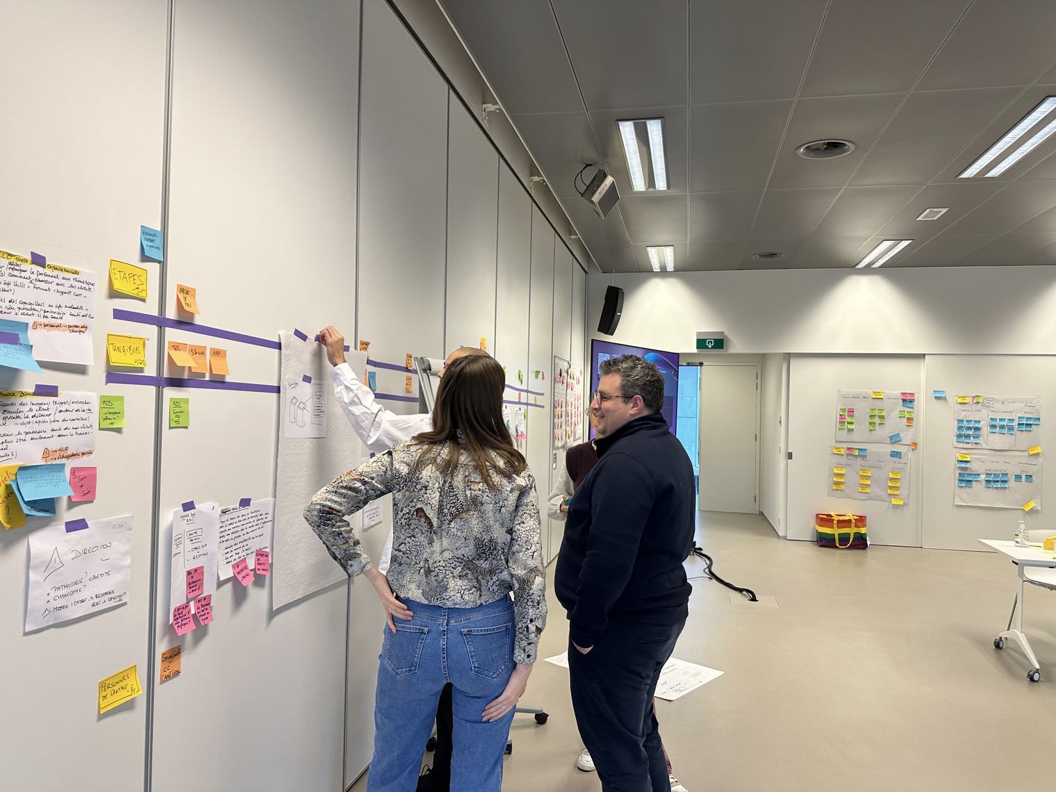 Trois personnes travaillent debout face à un mur couvert de feuilles et de post-its, en atelier de cadrage