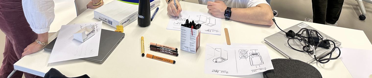 Table de travail en atelier : croquis d'un objet physique dessinés au marqueur, à côté d'ordinateurs portables